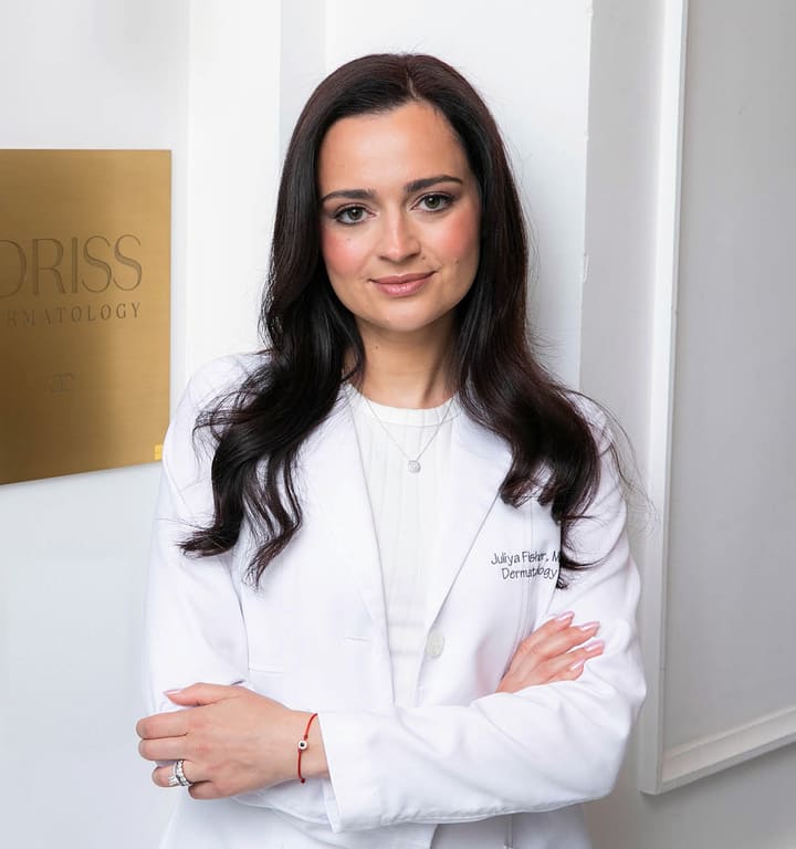 Dr. Juliya Fisher in her white coat beside the Idriss Dermatology plaque