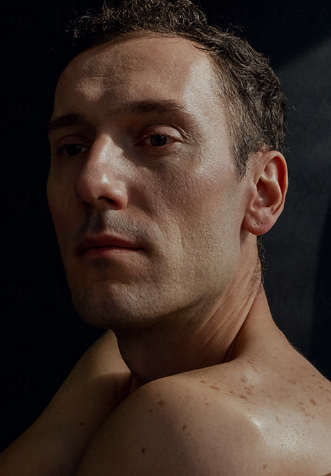 A man with short dark hair glancing over his bare shoulder, in low light